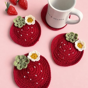 以下が含まれることがあります： 白い刺繍の種模様、緑のかぎ針編みの葉、黄色い中心の白い花があしらわれた、3枚の赤い円形のかぎ針編みコースター。Dean & Delucaの文字が入った白い陶器のマグカップが、4枚目の同じコースターの上に置かれています。