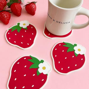 以下が含まれることがあります： 「DEAN & DELUCA」とプリントされた白いセラミックマグが、赤いイチゴ型のコースターの上に置かれています。さらに3つのイチゴコースターといくつかの人工イチゴがピンク色の表面に散らばっています。