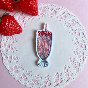 以下が含まれることがあります： ストローとホイップクリームが付いたピンク色のイチゴミルクシェイクの刺繍パッチ。パッチは白いペーパーコースターの上にあり、背景には3つの赤いイチゴがあります。背景は薄いピンク色です。