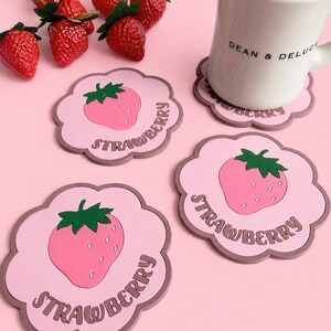 以下が含まれることがあります： 緑のヘタが付いたピンクのイチゴと茶色の「STRAWBERRY」の文字が中央に描かれた、スカラップ型のライトピンクのコースター3枚。「DEAN & DELUCA」の文字が入った白いセラミック製のマグカップがコースターの上に置かれ、その近くには新鮮なイチゴがいくつか並んでいる。