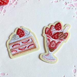 以下が含まれることがあります： イチゴケーキのスライスとイチゴパフェの刺繍パッチ。ケーキのスライスは白いフロスティングと赤いイチゴのフィリングが付いています。パフェは、イチゴとチョコレートの層になっています。