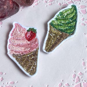 以下が含まれることがあります： 2つの刺繍入りアイスクリームコーンパッチ。1つは赤いイチゴが付いたピンク色のソフトクリーム、もう1つは緑色のソフトクリームです。どちらも金色のコーンのディテールと白い縁取りがあります。パッチはピンク色の背景にあります。