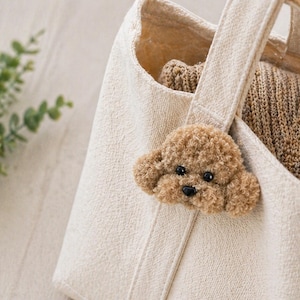 May include: A cream canvas tote bag with a fluffy, brown poodle dog applique on the handle. The bag contains tan-coloured knitwear. The dog has black eyes and a black nose.