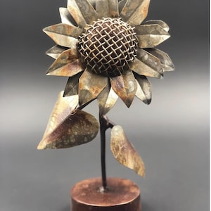 May include: A metal sunflower sculpture with a rustic finish. The sunflower has a wire mesh center and two leaves. The sculpture is mounted on a wooden base.