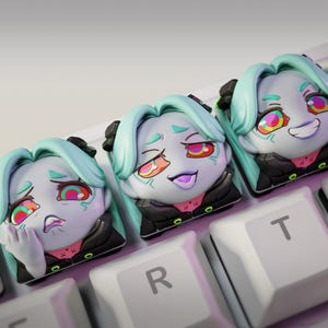 May include: Three custom keycaps featuring a stylized anime character with teal hair and expressive faces. The keycaps are on a white keyboard, with the "nom nom figures" logo visible in the corner. Each keycap has a different facial expression.