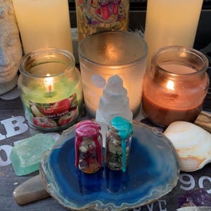 Op de afbeelding: Een stilleven met twee kleine glazen potjes met kleurrijke wasdruppels, rustend op een blauwe geode schijf. De potjes worden omringd door brandende kaarsen in glazen containers, een witte kristal en andere decoratieve items. De scène roept een spiritueel thema op.
