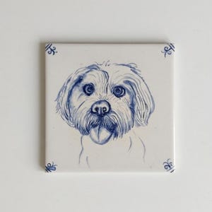 May include: A square white ceramic tile featuring a hand-painted blue ink illustration of a shaggy-haired dog with its tongue out. The corners of the tile are decorated with small blue scrollwork patterns, creating a clean and simple aesthetic.