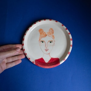 May include: A white ceramic plate with red and white stripes around the edge. A hand-painted portrait of a person with red hair and a red shirt is in the center of the plate.
