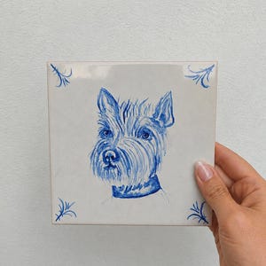 May include: A square white ceramic tile featuring a blue hand-painted illustration of a terrier dog wearing a collar. The artwork is centered on the tile, with small blue decorative flourishes in each of the four corners of the tile surface.