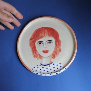 May include: Hand-painted ceramic plate featuring a portrait of a woman with red hair, blue eyes, and a red lip. The woman is wearing a blue and white polka dot top. The plate has a cream-colored surface with a peach-colored rim.