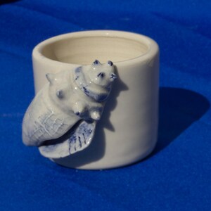 Puede incluir: Un pequeño recipiente cilíndrico de cerámica con esmalte blanco. Una figura de oso esculpida en azul y blanco está adherida al lateral. El oso está tumbado sobre una forma de concha azul y blanca. El recipiente tiene un borde liso y redondeado.