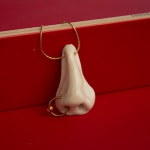 Op de afbeelding: Een grillige, crèmekleurige keramische neus-hanger met een goudkleurige oorring en een gouden ketting. De neus staat tegen een levendige rode achtergrond, wat een opvallend contrast creëert. De hanger is een uniek sieraad.