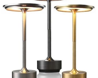 May include: Three modern table lamps in silver, black, and gold. Each lamp has a round, illuminated top, a textured stem, and a circular base. The lamps are designed for ambient lighting and decorative purposes.