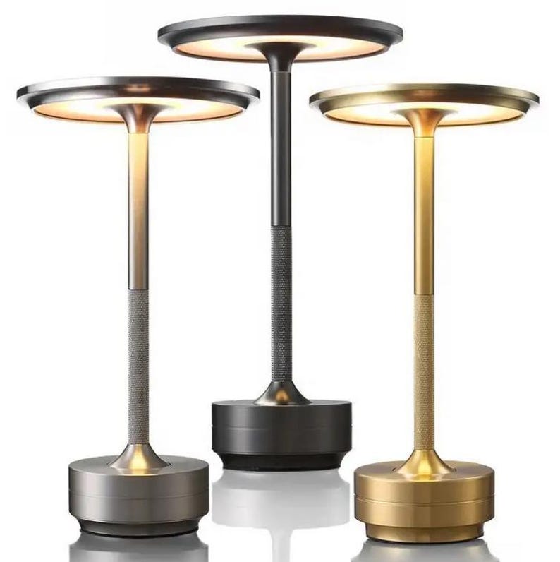 May include: Three table lamps with different colored bases. The lamps have a round, flat top with a light source in the center. The bases are made of metal and have a textured finish. The lamps are in a variety of colors, including gold, silver, and black.