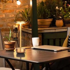 May include: A gold table lamp with a round, white shade. The lamp is on a wooden table with a laptop, a mug, and a potted plant.