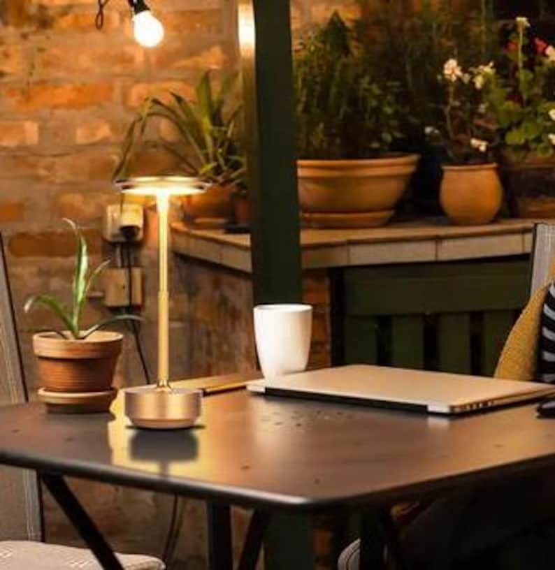 May include: A gold table lamp with a round, white shade. The lamp is on a wooden table with a laptop, a mug, and a potted plant.
