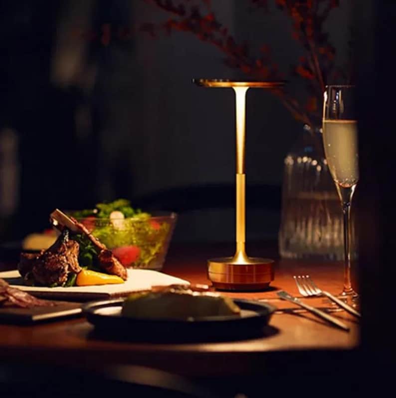 May include: A gold table lamp with a round base and a thin, vertical stem. The lamp has a circular shade that is slightly angled. The lamp is on a wooden table with a plate of food and a glass of wine.
