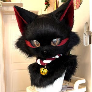May include: A black cat head with red accents around the eyes and ears. The cat has large, reflective eyes and a fluffy appearance. It wears a collar with a gold bell pendant. The cat is a costume or cosplay item.