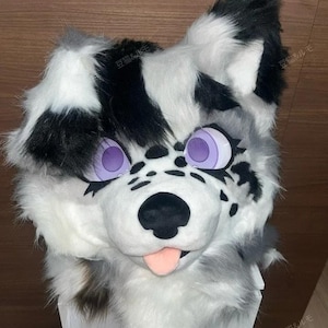 May include: A plush animal head mask featuring long white, black, and gray faux fur. The mask has large purple eyes with black eyelashes, a black nose, and a small pink tongue protruding from the mouth. The fur has black spots on the muzzle.