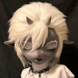 May include: A doll with a gray face, white hair, and small gray horns. The doll has large white eyes and pointed ears. It wears a white shirt and a necklace with a chain.