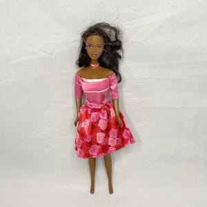 May include: A doll with dark brown hair and brown skin wearing a pink off-the-shoulder top with white trim and a red skirt featuring a pattern of pink and red roses. The doll has a small white necklace with a pink center accent around its neck.