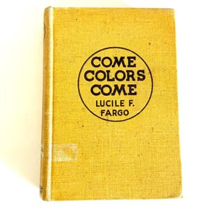Op de afbeelding: Een vintage boek met een mosterdgele kaft. De titel "COME COLORS COME" is gedrukt in een zwarte cirkel, met de naam van de auteur, Lucile F. Fargo, eronder. De rug van het boek is gebroken wit en de pagina's zijn verouderd.