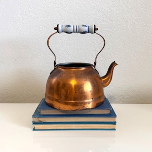 May include: A copper tea kettle with a curved spout and a metal handle featuring a white ceramic grip decorated with blue patterns. The kettle has a rounded body and sits atop a stack of two blue books with visible light-colored pages.