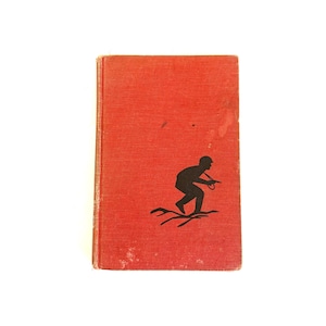 May include: A rectangular hardcover book with a textured red fabric cover. The front cover features a black silhouette graphic of a person in a helmet crouching or running over uneven ground. The cover shows signs of wear with light surface marks.