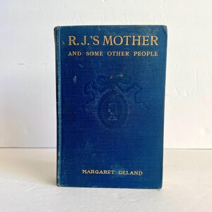 Pode incluir: Um livro encadernado em azul intitulado "R.J.'s Mother and Some Other People" de Margaret Deland.