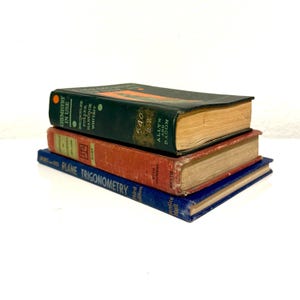 May include: Three vintage books stacked on top of each other. The top book is dark green with the title "CHEMISTRY IN USE". The middle book is red with the title "HART". The bottom book is blue with the title "PLANE TRIGONOMETRY".