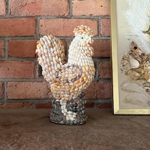 May include: A decorative rooster figurine crafted entirely from various small seashells. The body features a mix of white, tan, and brown shells, while the base is covered in darker, greyish-brown shells. The rooster stands in a profile position.