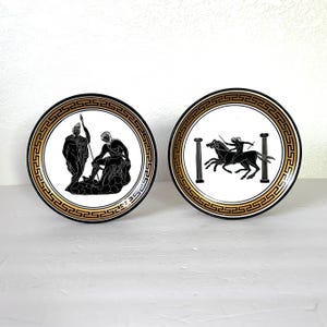 May include: Two decorative ceramic plates featuring black and white Greek mythology scenes. The plates have a gold border with a repeating Greek key pattern.
