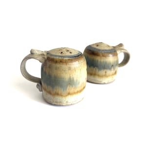 May include: Two ceramic salt and pepper shakers featuring a cylindrical body with a handle, a rounded top with small holes, and a glaze finish in blended shades of beige, tan, and muted blue, creating a layered, earthy appearance on each piece.