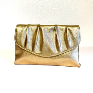 May include: A gold clutch with a pleated top flap. The rectangular bag has a metallic sheen and a curved bottom edge. The clutch is a solid gold color and appears to be made of a soft material.