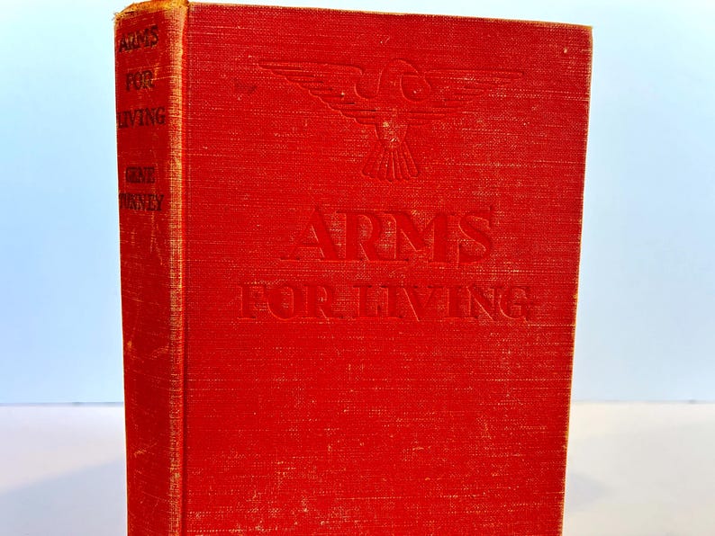 May include: A red hardback book with the title "Arms for Living" embossed on the front cover. The book features a stylised eagle design above the title.