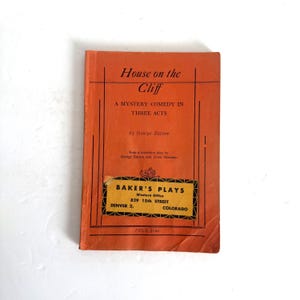 Könnte beinhalten: Ein orangefarbenes Taschenbuch-Theaterstück mit dem Titel House on the Cliff, A Mystery Comedy in Three Acts von George Batson. Das Cover zeigt schwarzen Text und ein gelbes rechteckiges Etikett für Baker's Plays, Western Office, 829 15th Street, Denver 2, Colorado.
