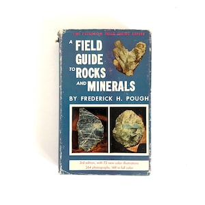 May include: A vintage field guide titled "A Field Guide to Rocks and Minerals" by Frederick H. Pough. The cover is blue with the title in white and green. The book features illustrations of rocks and minerals, with text indicating it's the 3rd edition with 72 color illustrations.
