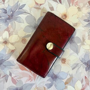 May include: A rectangular dark red leather wallet with a textured, marbled finish. The wallet features a vertical strap closure secured by a polished, circular gold-tone metal snap button centered on the front flap of the accessory.