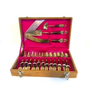 Può includere: Un set di posate vintage in una valigetta di legno aperta con interno rosa acceso. Il set include forchette, cucchiai, coltelli e utensili da portata. I manici sono marrone scuro con dettagli metallici decorativi. La valigetta è aperta, rivelando i pezzi ben disposti.