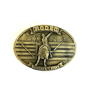 May include: Gold-coloured, oval belt buckle depicting a rodeo scene. It shows a cowboy on a bull, with the word "RODEO" above and "CANADA'S #1 SPORT" below. The buckle has a detailed, embossed design, typical of a vintage accessory.