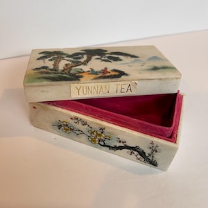 May include: A white rectangular box with a painted scene of a mountain landscape and trees. The box has a pink velvet interior and a gold label that reads "YUNNAN TEA".