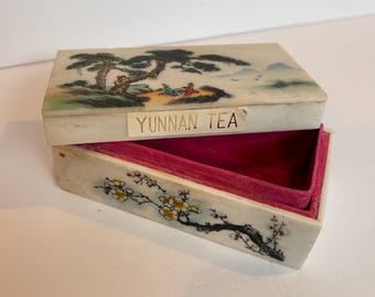 May include: A white rectangular box with a painted scene of a mountain landscape and trees. The box has a pink velvet interior and a gold label that reads "YUNNAN TEA".