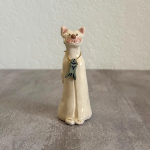May include: A tall, slender ceramic cat figurine with a glossy off-white finish. The cat has a small pink nose and mouth, and wears a thin gold-colored chain around its neck featuring a small, dark gray, fish-shaped pendant hanging at the center.