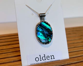 Pode incluir: Um colar de corrente de prata com um pingente que apresenta uma concha de abalone azul e verde engastada em uma moldura oval de prata. O pingente está em um cartão branco com o texto "olden" e um endereço de site.