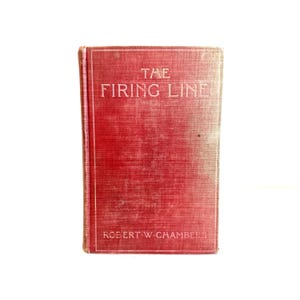 Pode incluir: Um livro antigo com capa vermelha, intitulado "The Firing Line" de Robert W. Chambers. A capa é vermelha desbotada, com o título e o nome do autor em letras brancas. O livro mostra sinais de desgaste.