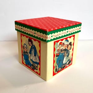 Puede incluir: Una caja de regalo cuadrada, roja y verde con fondo blanco y un patrón repetido de corazones rojos en la parte superior. La caja presenta ilustraciones de las muñecas Raggedy Ann y Andy en los lados. El texto "Raggedy Ann & Andy" está impreso en la caja.