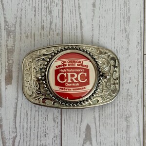 May include: An oval silver-tone metal belt buckle featuring an intricate embossed scrollwork border and a central circular emblem. The emblem has a red background with white text reading CRC CHEMICALS SUPER DIRT SERIES HIGH PERFORMANCE CRC.