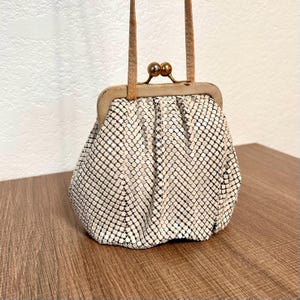 Könnte beinhalten: Eine Vintage-Handtasche aus weißem Netzstoff mit Kussverschluss und hellbraunem Griff. Die Handtasche hat ein zartes, strukturiertes Aussehen und eine kleine, abgerundete Form. Der Metallrahmen und der Verschluss sind goldfarben.