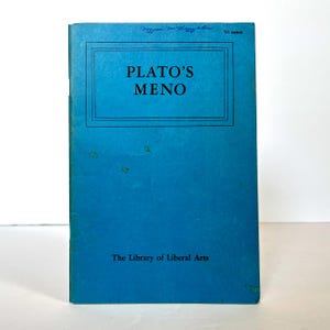 Op de afbeelding: Een blauw boek met de titel "PLATO'S MENO" in een zwart kader. Het boek maakt deel uit van de serie "The Library of Liberal Arts".