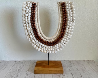 May include: A U-shaped decorative object featuring a dense arrangement of small white seashells and dark brown wooden beads on a cream-colored woven base. The piece is mounted on a thin black metal rod attached to a square wooden base.